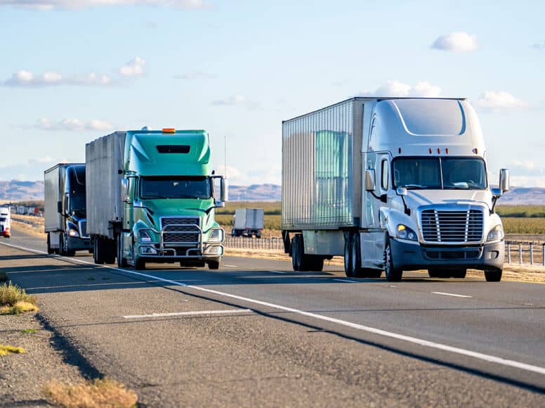 Truck Industries Two semi-trucks traveling on a multi-lane highway, with a white tractor-trailer in front and a green truck behind.