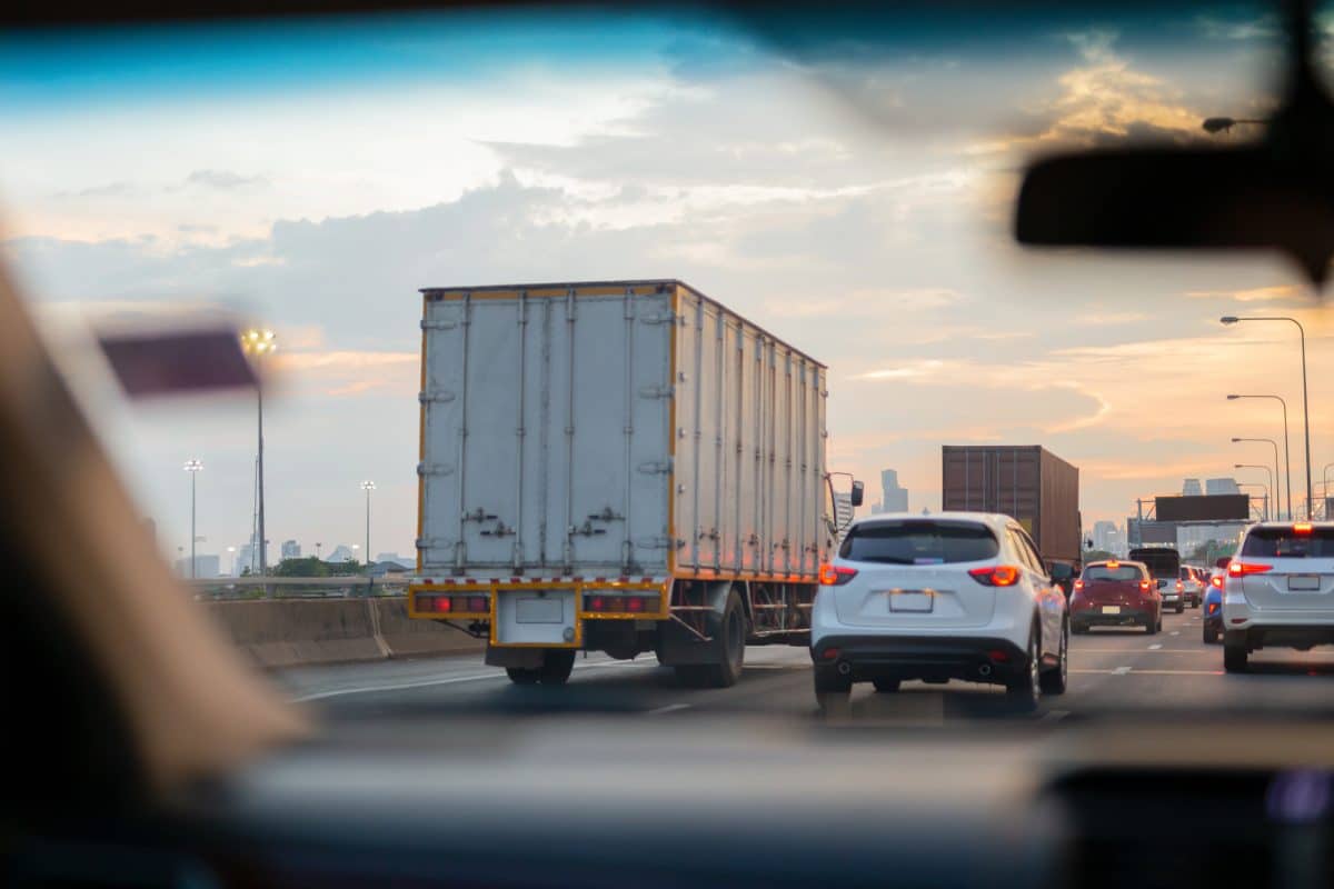Depositphotos_271949230_L View through a car windshield of a box truck and several vehicles traveling in heavy highway traffic at sunset.