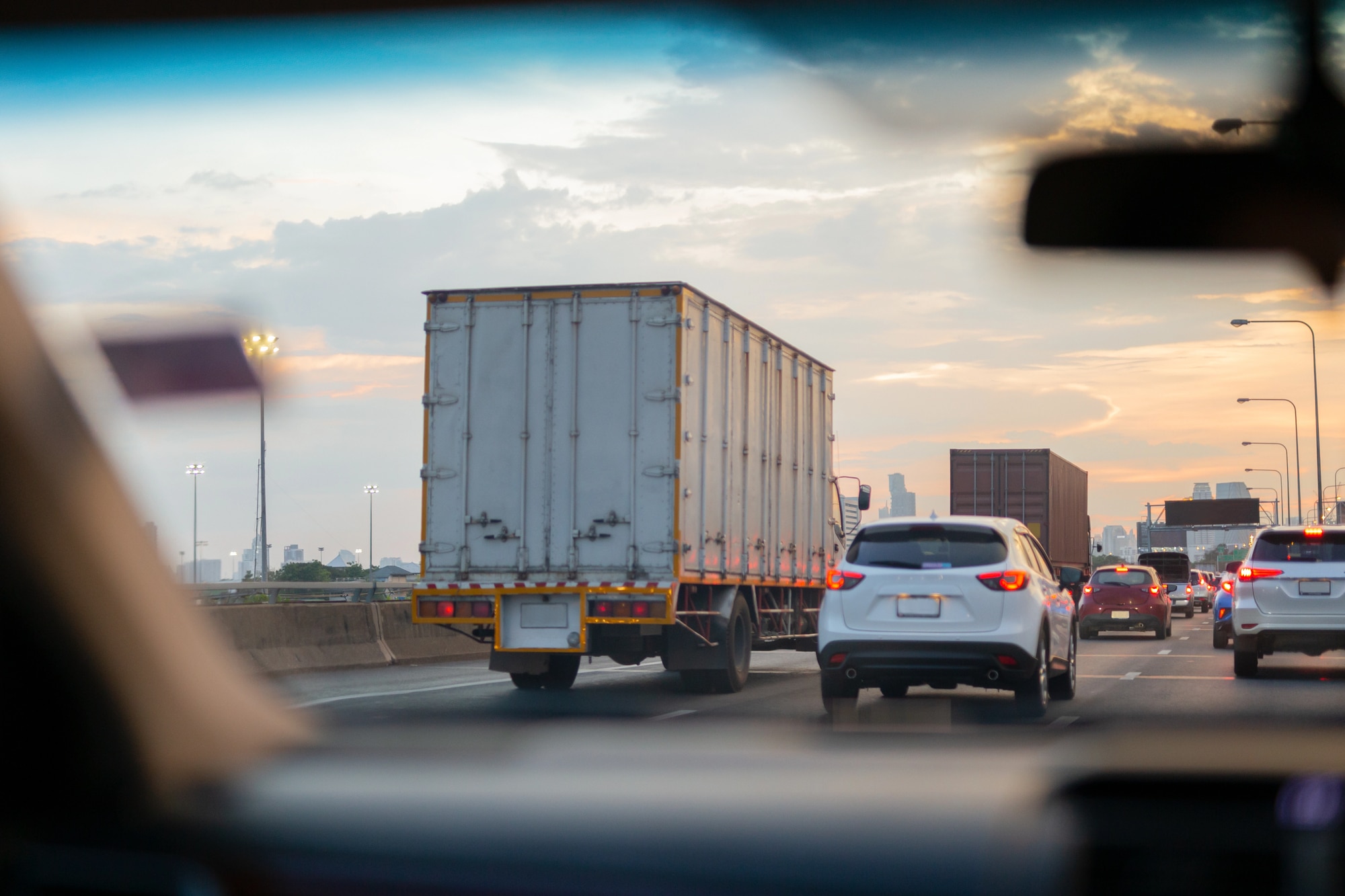 View through a car windshield of a box truck and several vehicles traveling in heavy highway traffic at sunset.