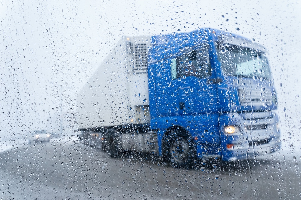 Blue semi-truck driving on a highway in heavy rain, viewed through a windshield covered in water droplets.